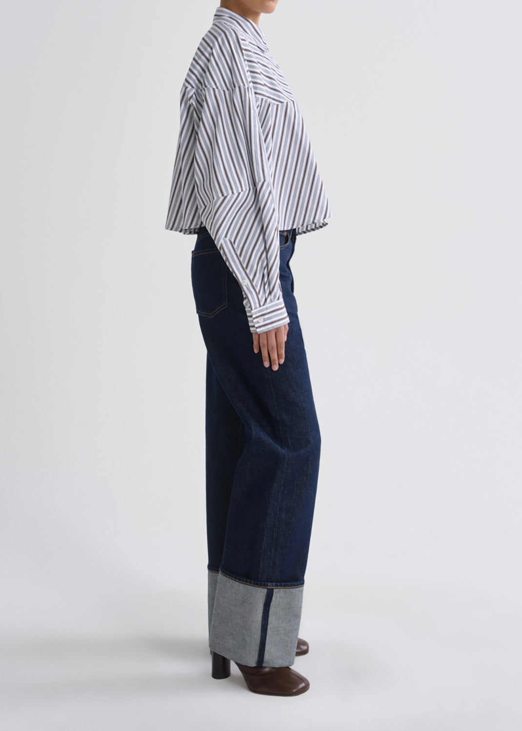 A model shown from the side profile wearing a white long-sleeve button-up shirt with thin dark vertical stripes, paired with dark blue wide-leg jeans featuring a cuffed hem showing a heather gray lining, and brown shoes, against a grey background.