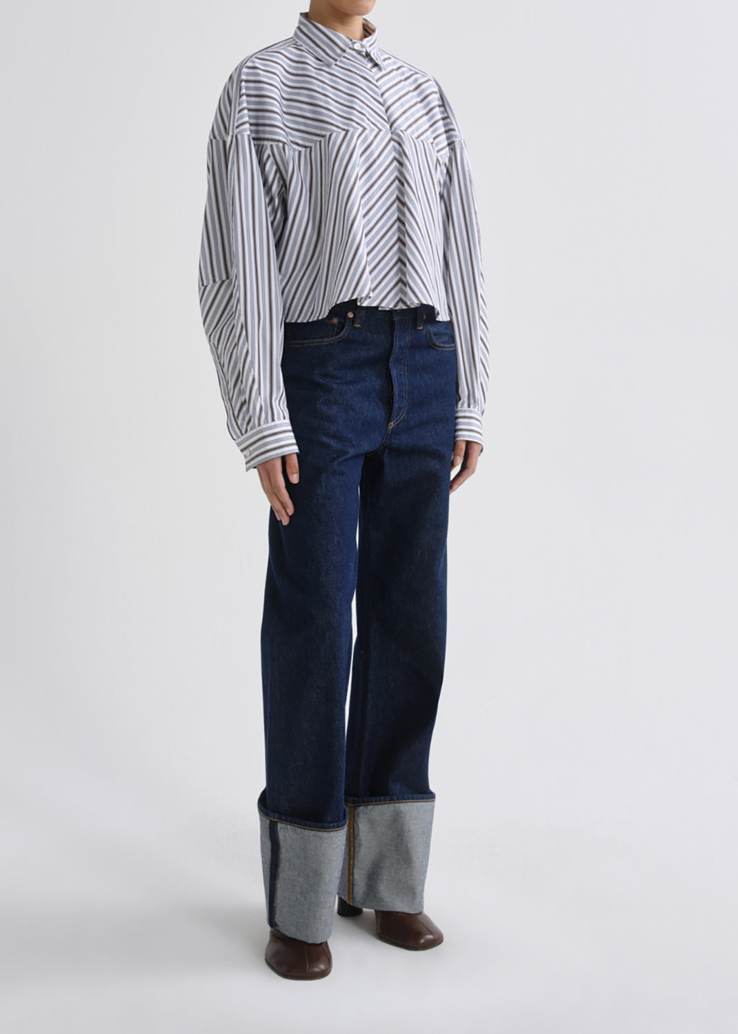 A model wearing a long-sleeve button-up shirt with thin vertical black and white stripes, paired with dark blue wide-leg jeans featuring a contrasting light gray cuffed hem and brown leather shoes, shot against a grey background.