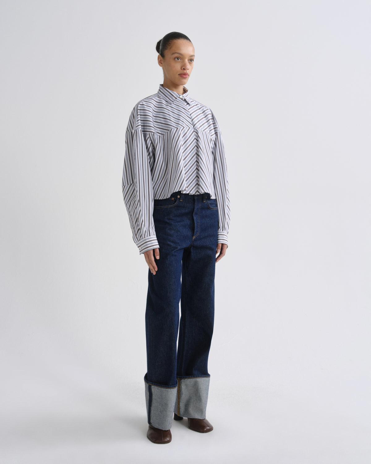 A model stands against a light gray studio background wearing a white long-sleeve button-up shirt with thin dark vertical stripes, dark blue wide-leg jeans with folded cuffs, and brown shoes.