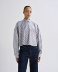 A model wearing a cropped, long-sleeve button-up shirt with diagonal navy and white stripes, paired with dark blue jeans, against a plain light gray studio background.