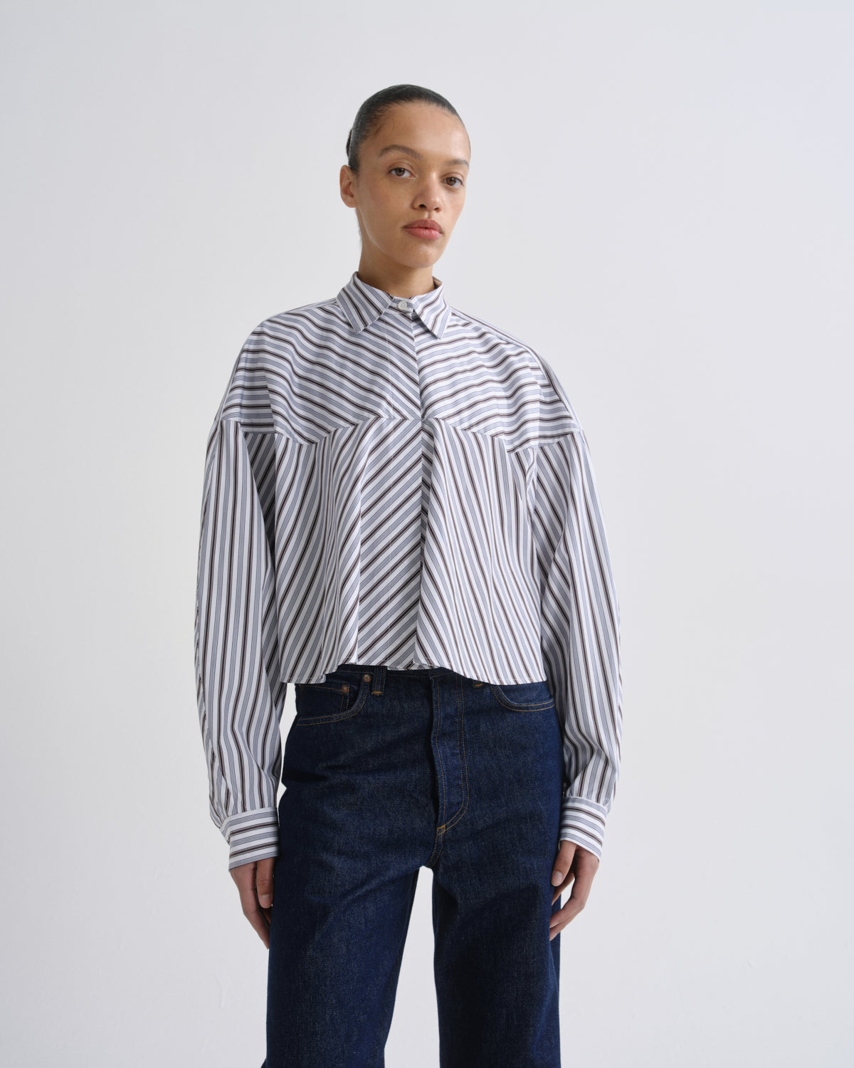 A model wearing a cropped, long-sleeve button-up shirt with diagonal navy and white stripes, paired with dark blue jeans, against a plain light gray studio background.