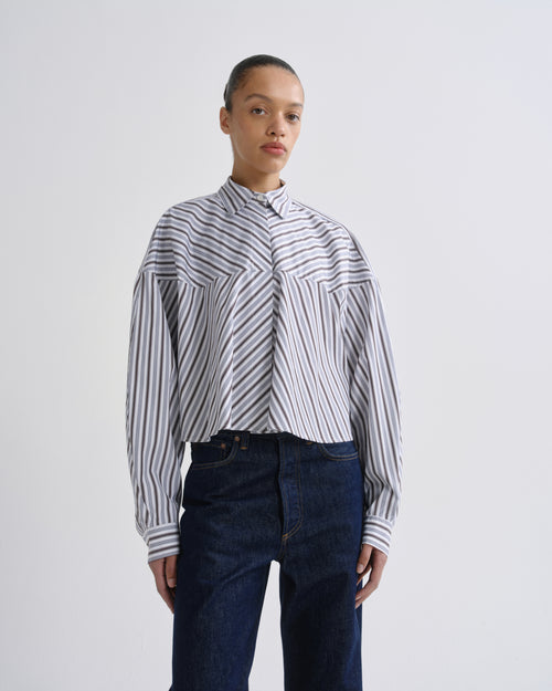 A model wearing a cropped, long-sleeve button-up shirt with diagonal navy and white stripes, paired with dark blue jeans, against a plain light gray studio background.