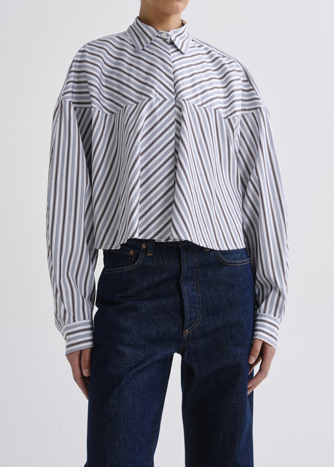 A model wearing a cropped, long-sleeve button-up shirt with vertical navy and white stripes, paired with dark blue jeans, against a grey background.