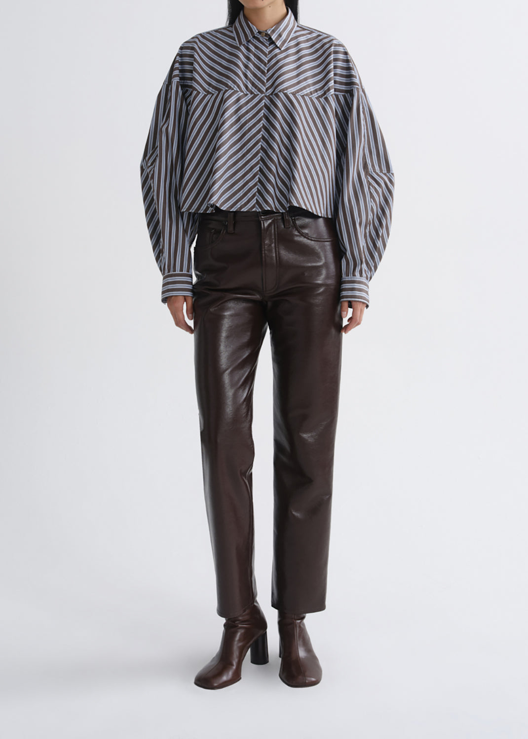 A model wearing a cropped, long-sleeve button-up shirt with diagonal grey and black stripes, paired with dark brown faux-leather trousers and matching brown heeled ankle boots, shot against a grey background.