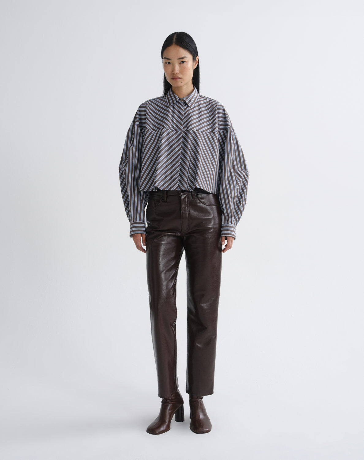A model stands against a light gray studio background wearing a black and white striped long-sleeve shirt with voluminous sleeves, dark brown faux-leather pants, and dark brown ankle boots.