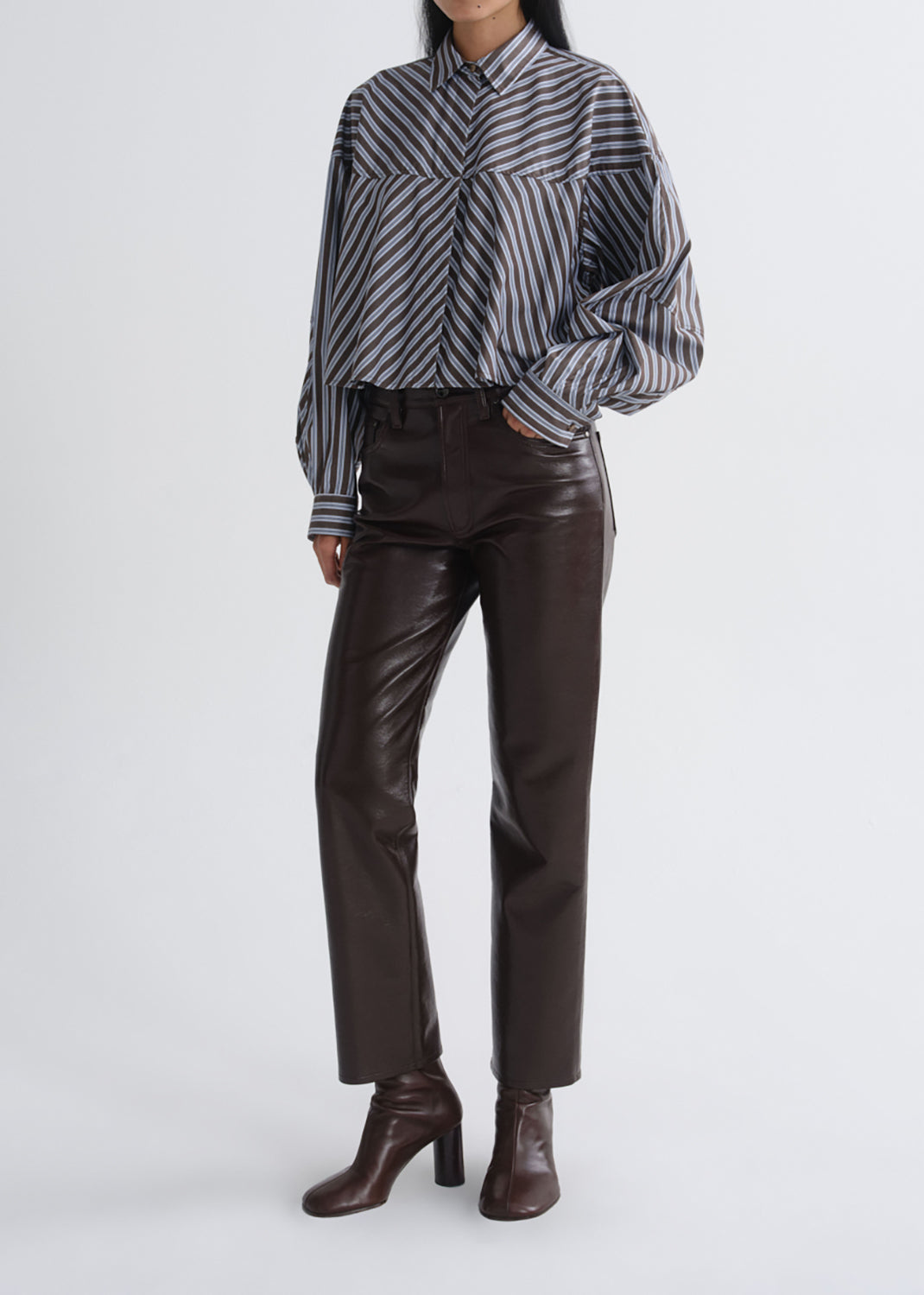 A model wearing a black and white striped long-sleeve shirt, dark brown faux-leather straight-leg pants, and dark brown heeled ankle boots, photographed against a grey background.