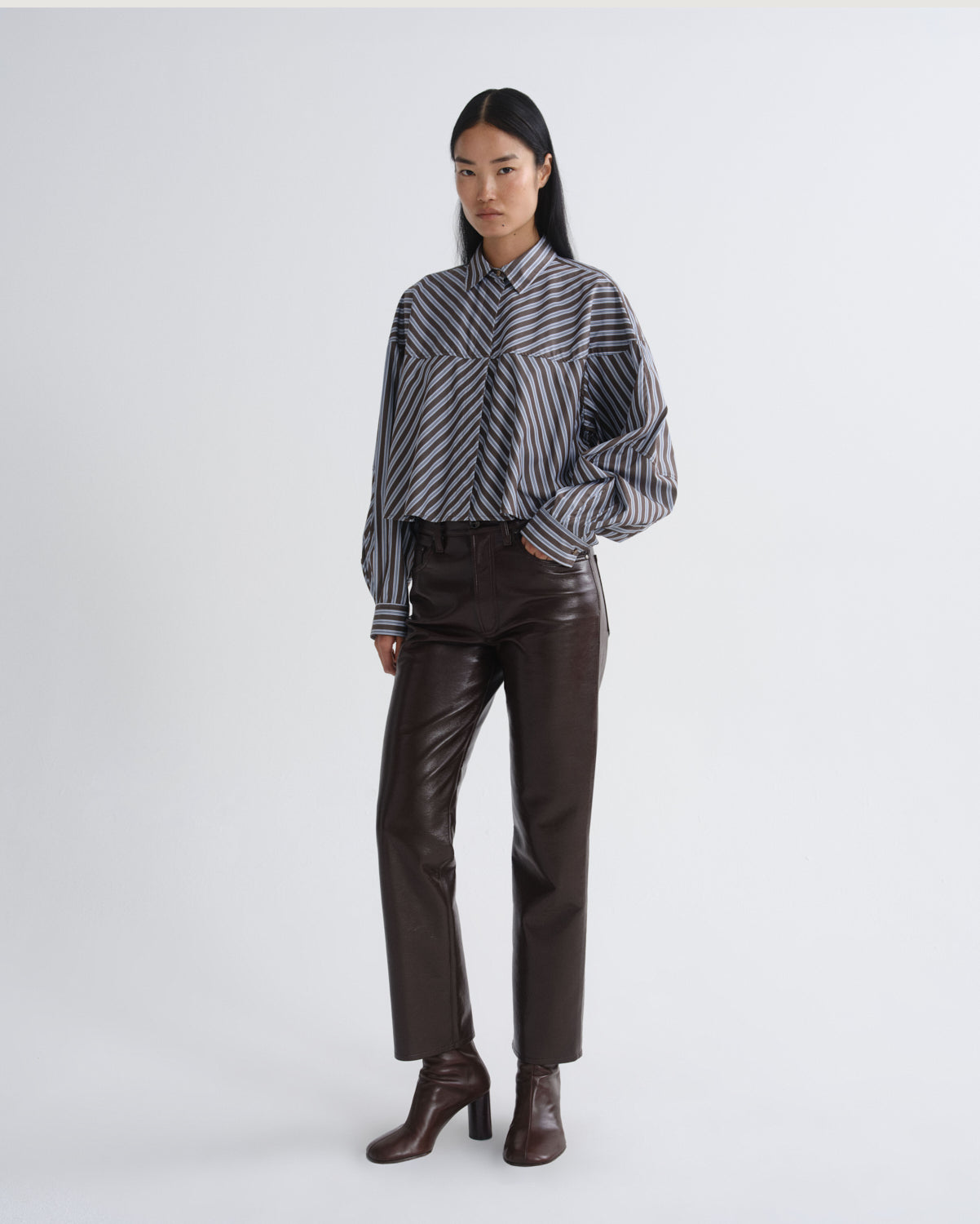 A model stands against a light gray studio background wearing a black and white vertically striped button-down shirt with voluminous sleeves, dark brown faux-leather trousers, and dark brown heeled ankle boots.
