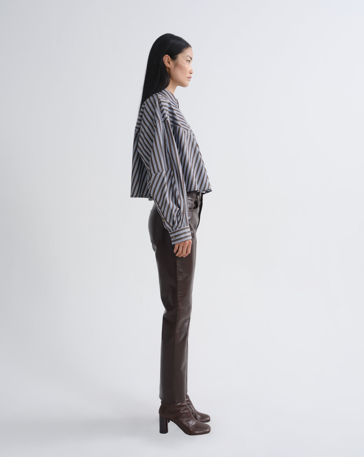 A woman stands in profile wearing a black and white vertically striped long-sleeve blouse, dark brown leather pants, and dark brown heeled ankle boots against a light gray studio background.
