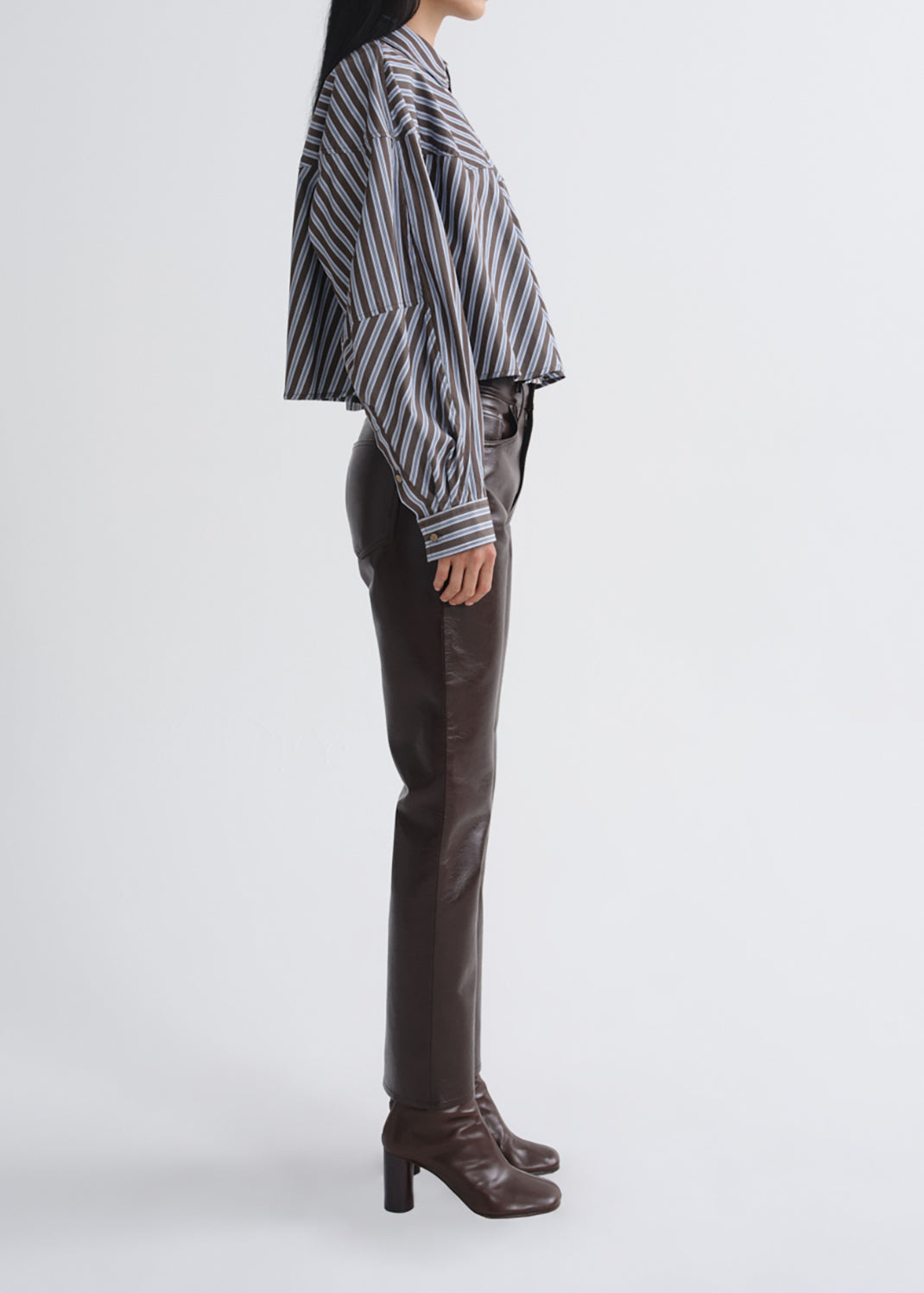 A model shown in profile wearing a cropped black and grey striped long-sleeve shirt, dark brown leather pants, and dark brown heeled ankle boots, set against a grey background.