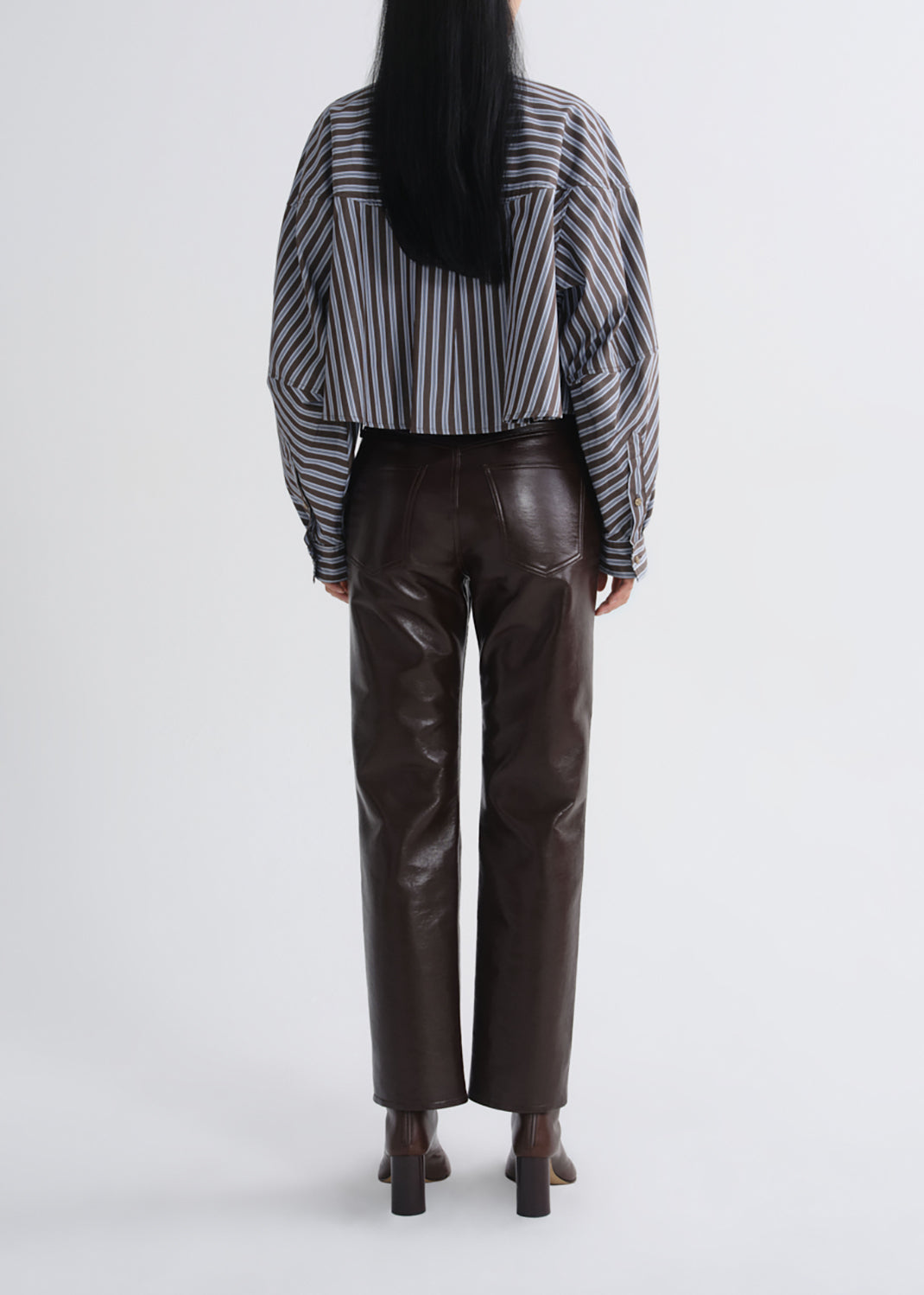 A model shown from the back wearing a black and white vertically striped cropped shirt, dark brown faux-leather pants, and dark brown block-heel boots against a grey background.