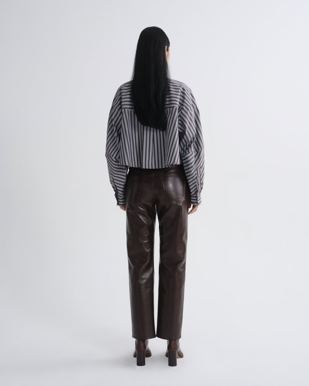 A model viewed from the back wearing a black and white vertically striped long-sleeve shirt and dark brown faux-leather trousers, set against a plain light gray studio background.