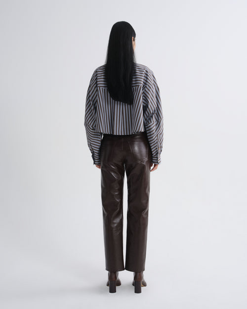 A model viewed from the back wearing a black and white vertically striped long-sleeve shirt and dark brown faux-leather trousers, set against a plain light gray studio background.