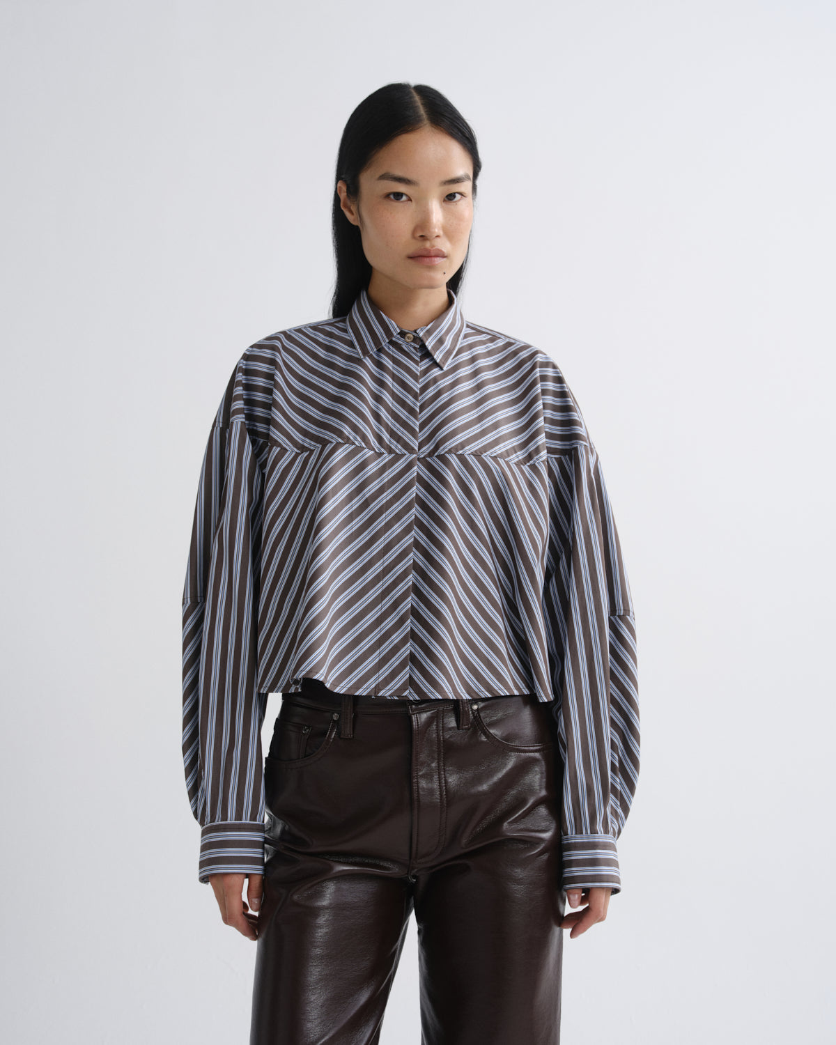 A model wearing a cropped, long-sleeve button-up shirt with diagonal black, gray, and white stripes, paired with dark brown faux-leather pants, against a light gray studio background.
