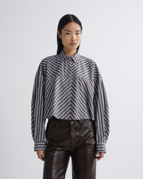 A model wearing a cropped, long-sleeve button-up shirt with diagonal black, gray, and white stripes, paired with dark brown faux-leather pants, against a light gray studio background.