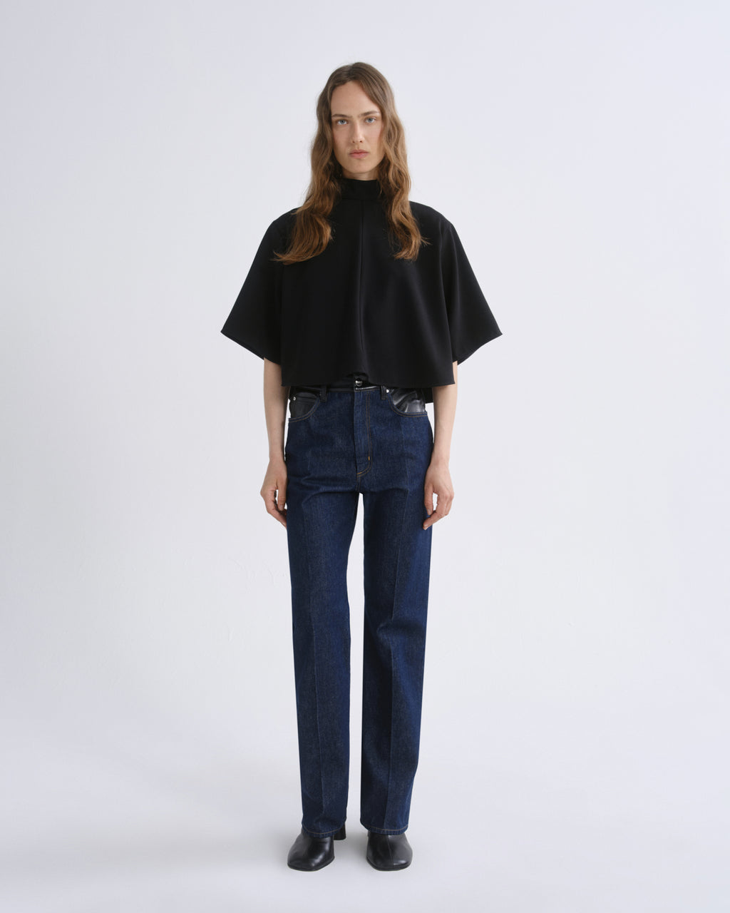 A woman stands against a grey background wearing a black short-sleeved mock-neck cape top, dark blue straight-leg jeans, and black shoes.