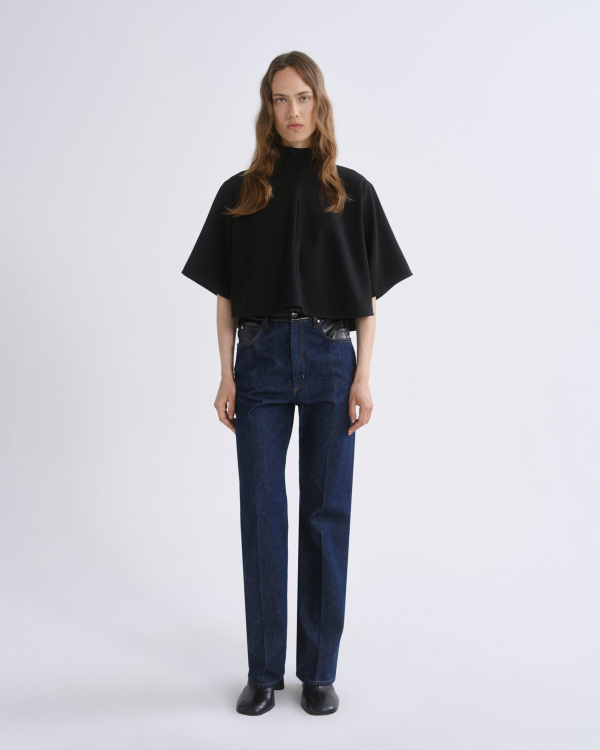 A woman stands against a grey background wearing a black short-sleeved mock-neck cape top, dark blue straight-leg jeans, and black shoes.
