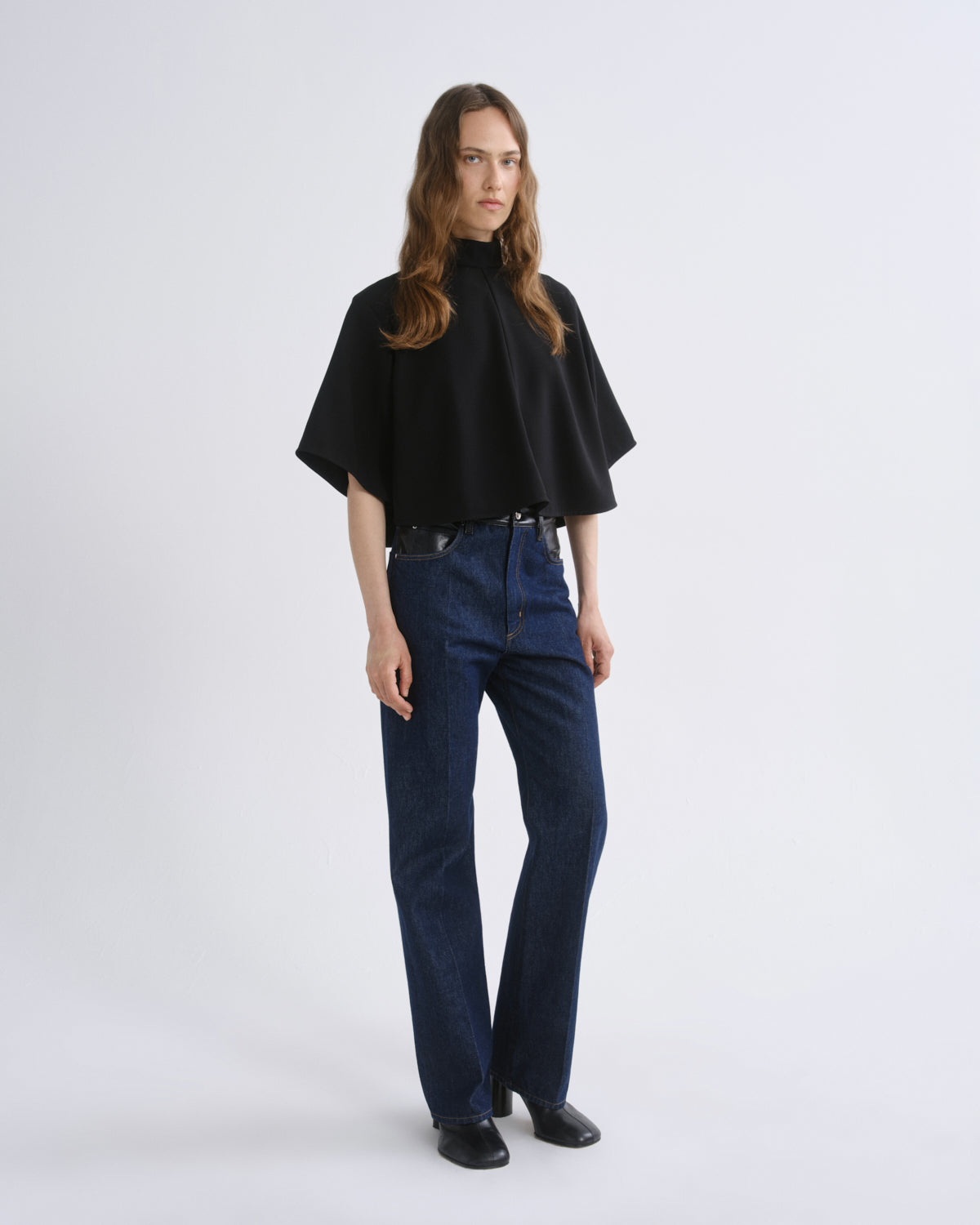 A woman modeling a black short-sleeved mock-neck cape top paired with dark blue jeans and black shoes, standing against a grey background.
