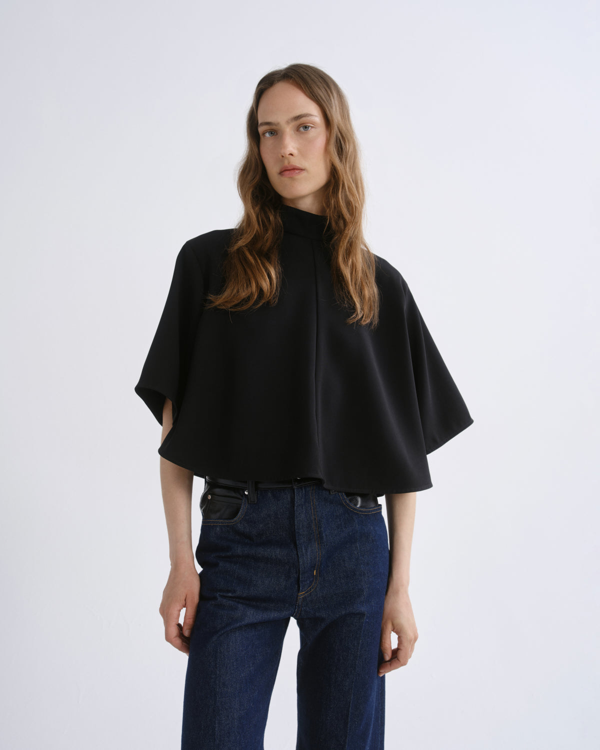 A woman modeling a black high-neck cape-style top with wide short sleeves, paired with dark blue jeans, against a grey background.