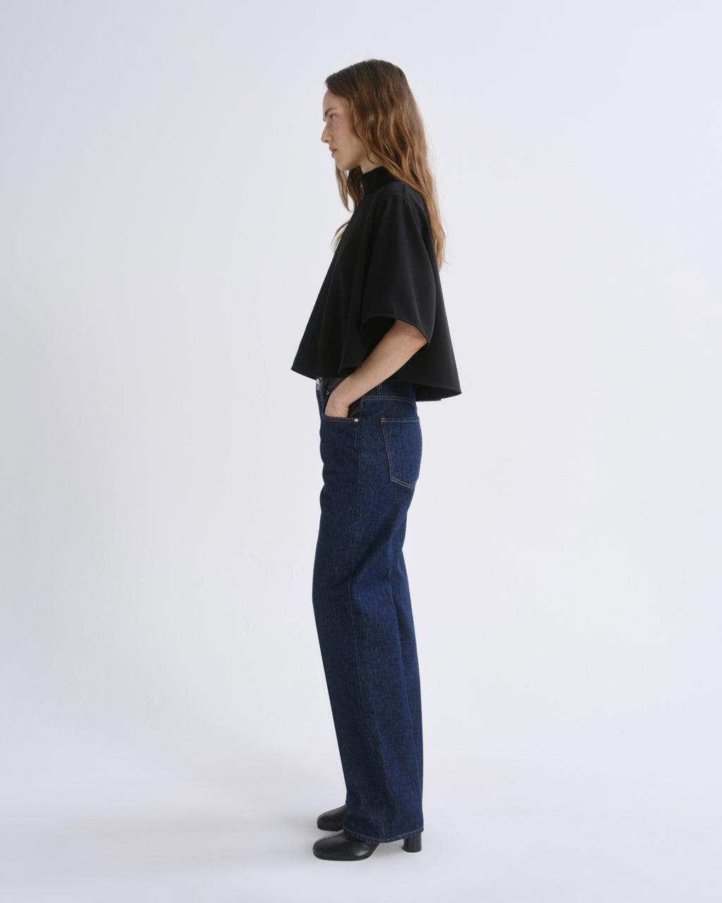 A woman standing in profile wearing a black cropped short-sleeve top, dark blue wide-leg jeans, and black shoes against a grey background.
