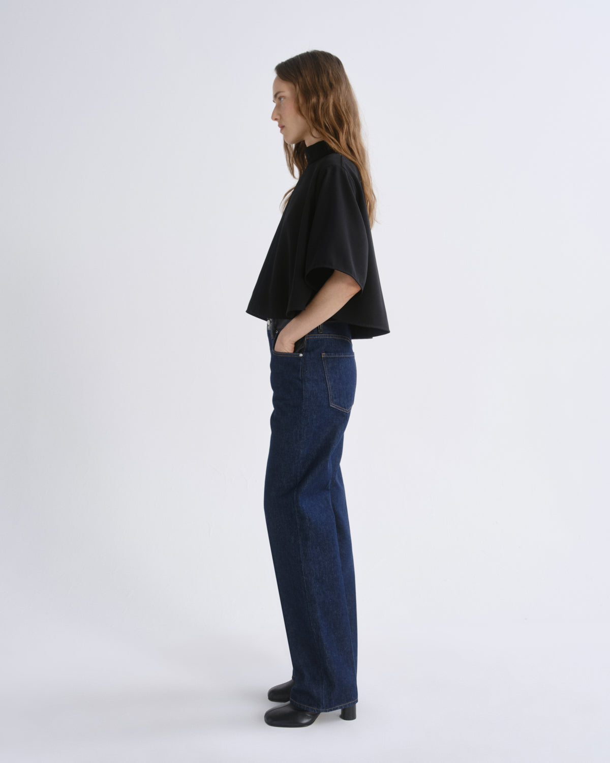 A woman standing in profile wearing a black cropped short-sleeve top, dark blue wide-leg jeans, and black shoes against a grey background.
