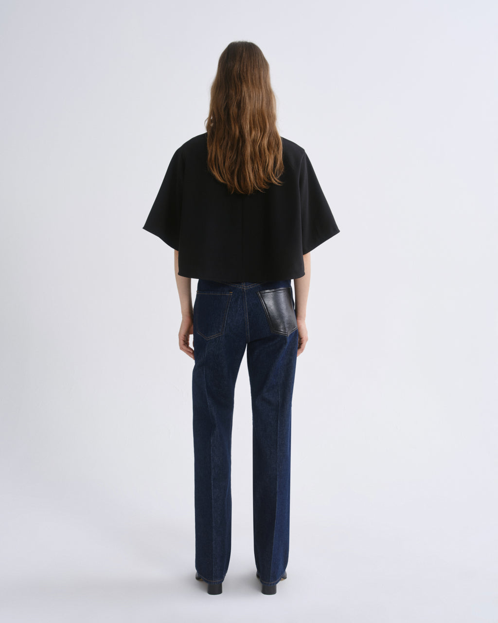 A model viewed from the back wearing a black short-sleeved cropped top and dark blue straight-leg jeans with a glossy black leather patch detail on the back pocket, set against a grey background.