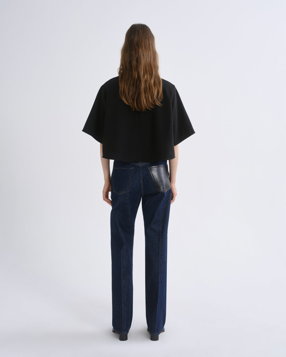 A model viewed from the back wearing a black short-sleeved cropped top and dark blue straight-leg jeans with a glossy black leather patch detail on the back pocket, set against a grey background.