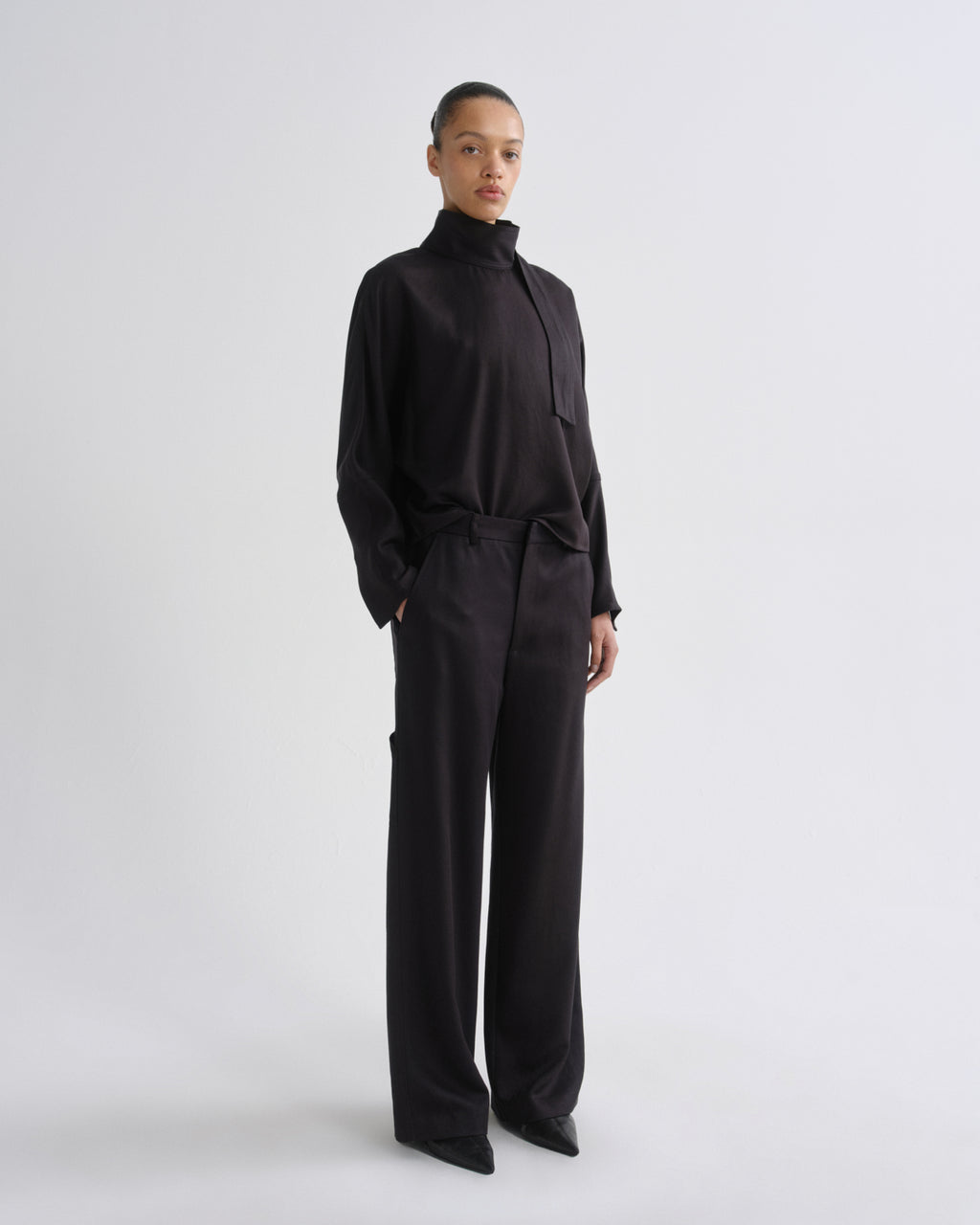 A model stands in a studio wearing a black long-sleeve mock-neck top and matching black wide-leg trousers against a light gray background.