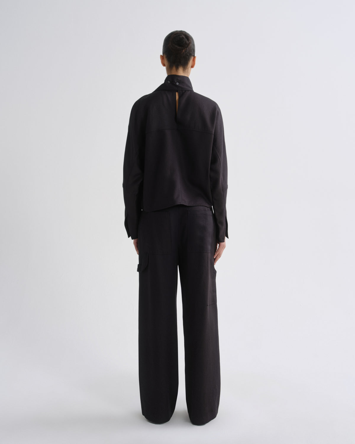 A model viewed from the back wearing a black long-sleeve top with a high mock neck and wide-leg black trousers, set against a plain light gray studio background.