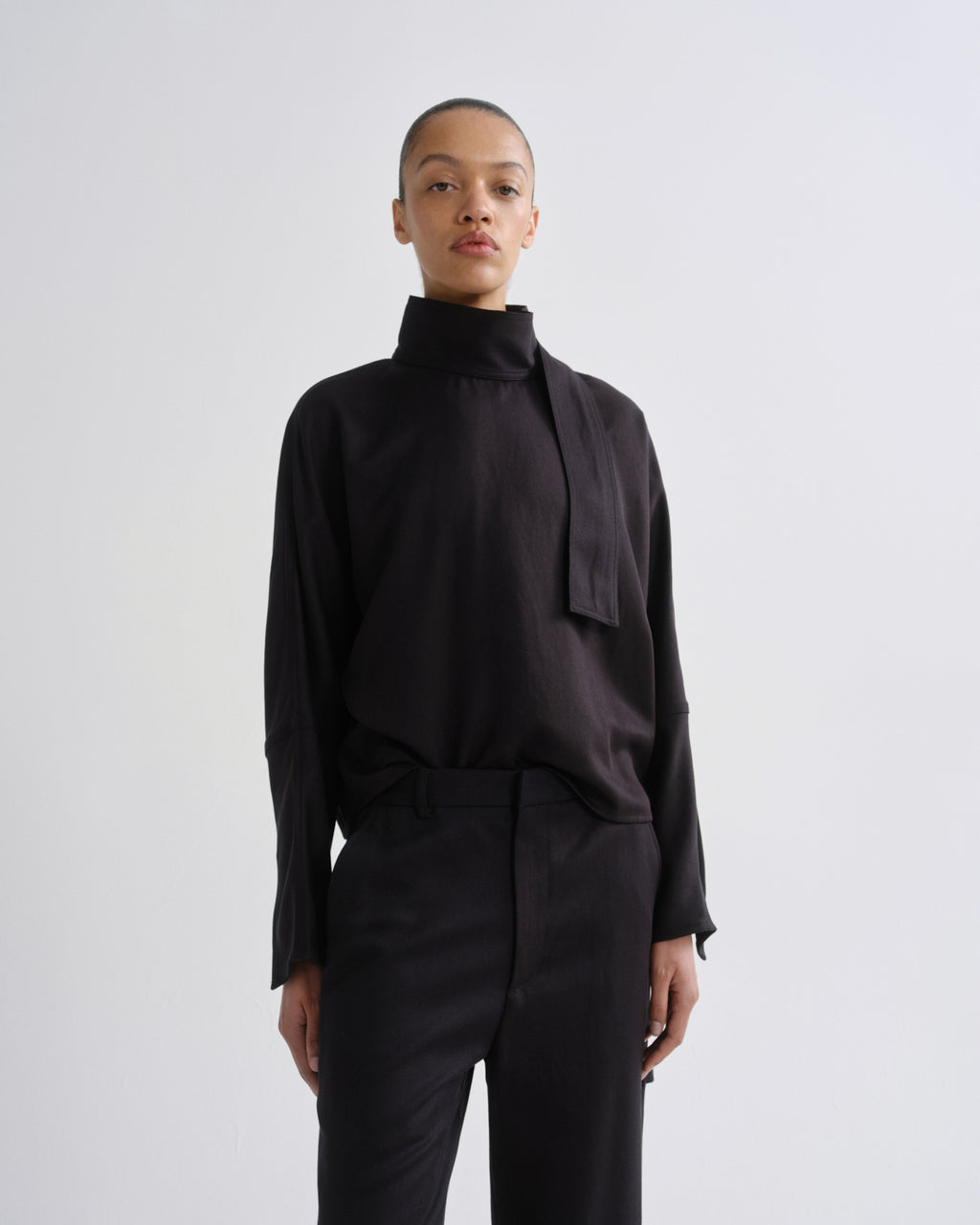 A model wearing a black long-sleeve high-neck blouse with a wide tie detail at the collar and black trousers, standing against a light gray studio background.