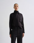 A model wearing a black long-sleeve high-neck blouse with a wide tie detail at the collar and black trousers, standing against a light gray studio background.