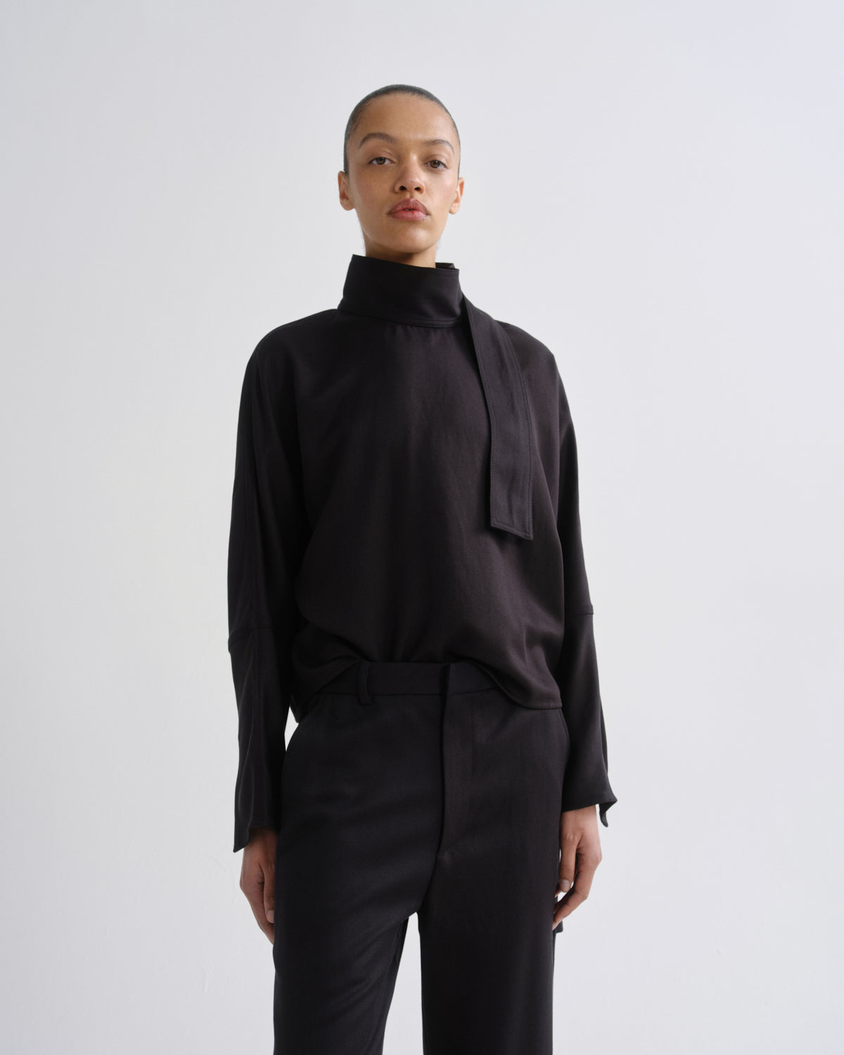 A model wearing a black long-sleeve high-neck blouse with a wide tie detail at the collar and black trousers, standing against a light gray studio background.