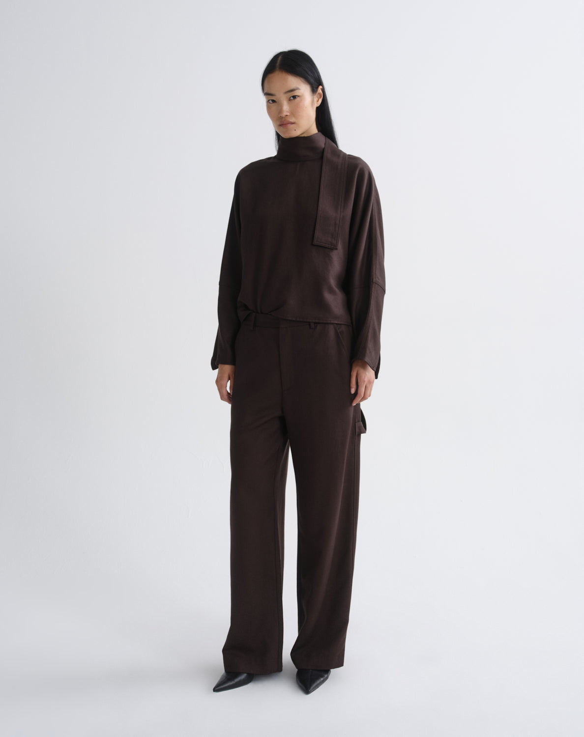 A model stands against a grey background wearing a dark brown high-neck long-sleeve top and matching wide-leg trousers.