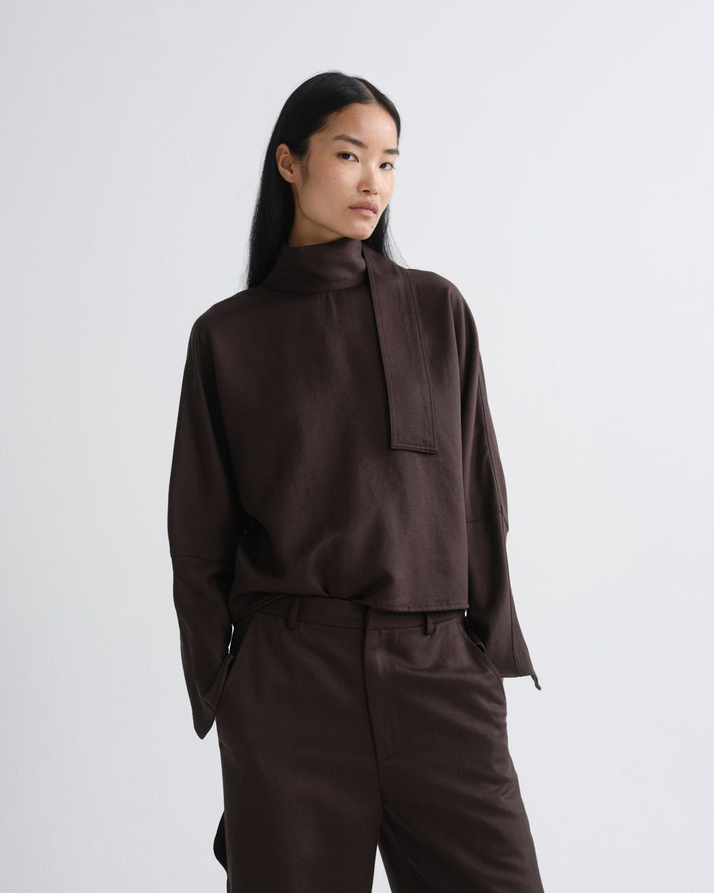A model wearing a dark brown high-neck long-sleeve top with a unique front tab detail and matching dark brown trousers, photographed against a grey background.