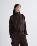 A model wearing a dark brown high-neck long-sleeve top with a unique front tab detail and matching dark brown trousers, photographed against a grey background.