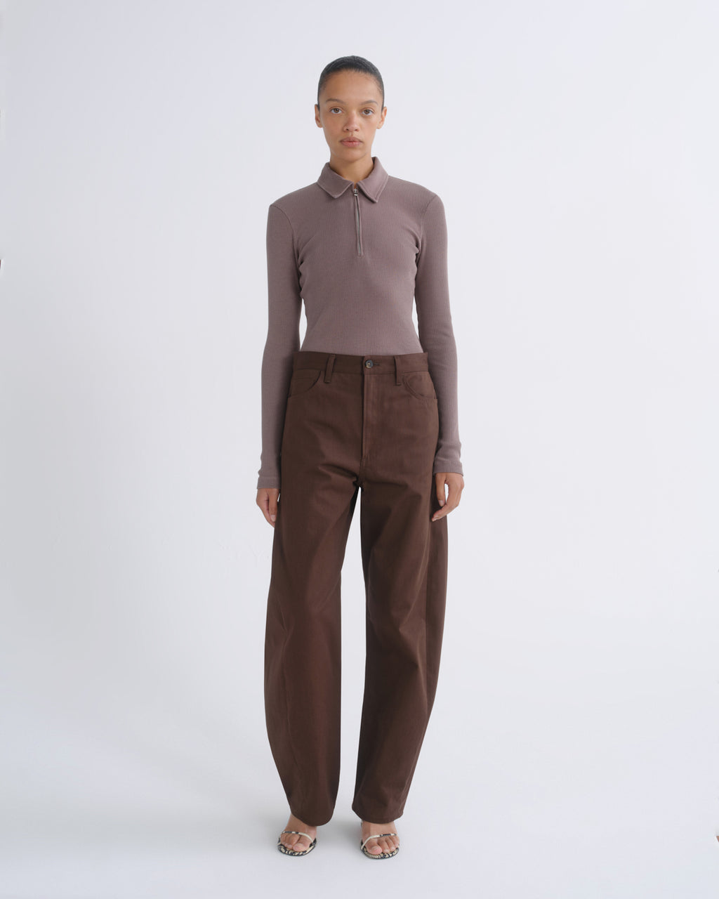 A model stands against a light gray studio background wearing a fitted long-sleeve polo shirt in muted taupe-brown and wide-leg brown trousers.