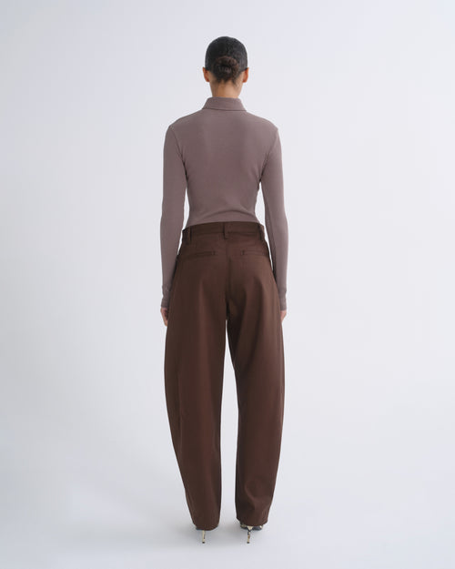 A model wearing a taupe long-sleeve polo-neck knit top and dark brown trousers, standing against a light gray studio background.