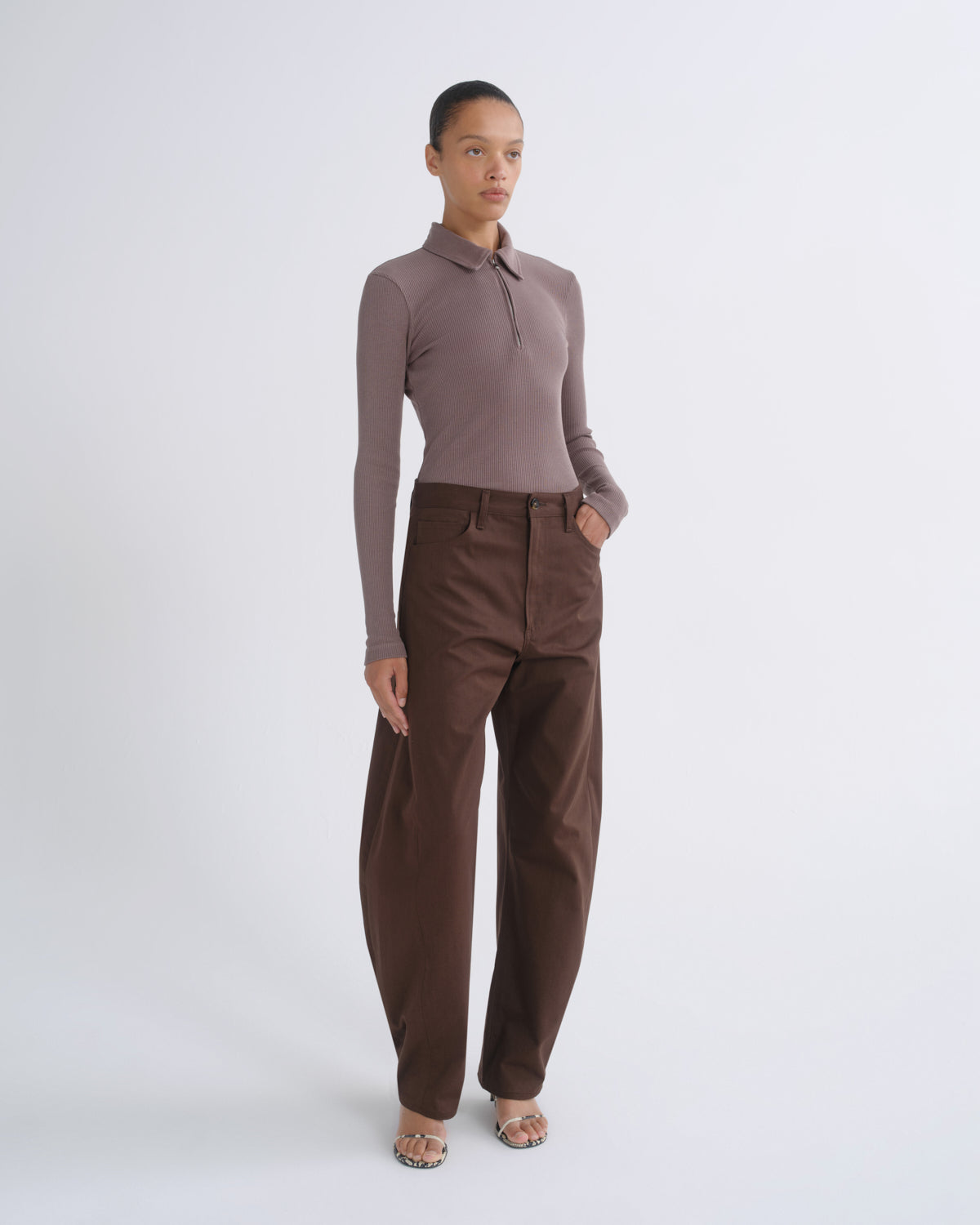 A model stands against a light gray studio background wearing a fitted long-sleeve polo top in taupe and high-waisted brown trousers.