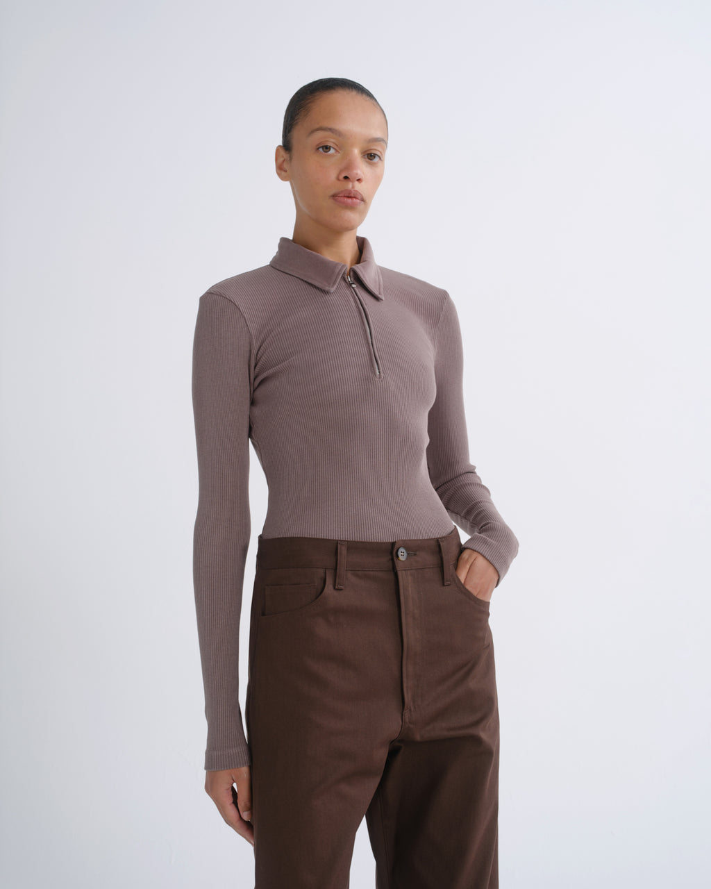 A model wearing a taupe long-sleeve polo-neck knit top and dark brown trousers, standing against a light gray studio background.