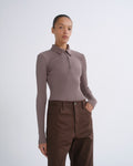 A model wearing a taupe long-sleeve polo-neck knit top and dark brown trousers, standing against a light gray studio background.