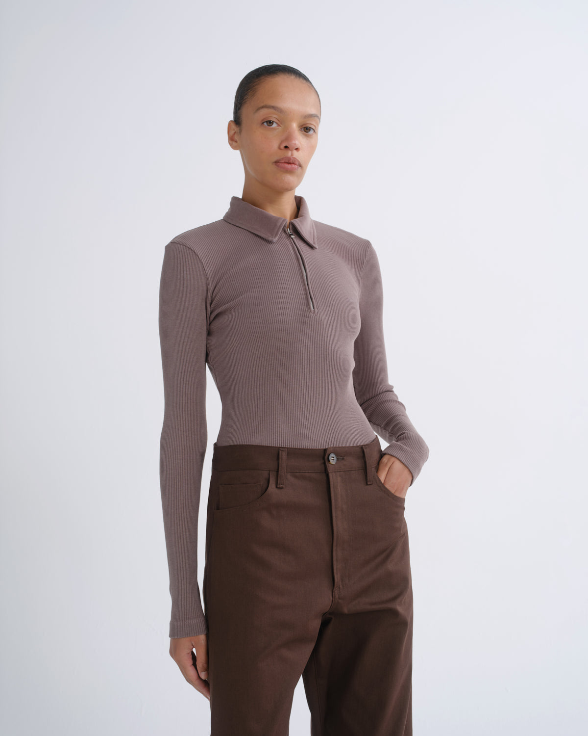 A model wearing a taupe long-sleeve polo-neck knit top and dark brown trousers, standing against a light gray studio background.