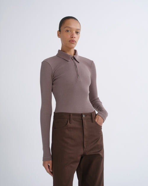 A model wearing a taupe long-sleeve polo-neck knit top and dark brown trousers, standing against a light gray studio background.