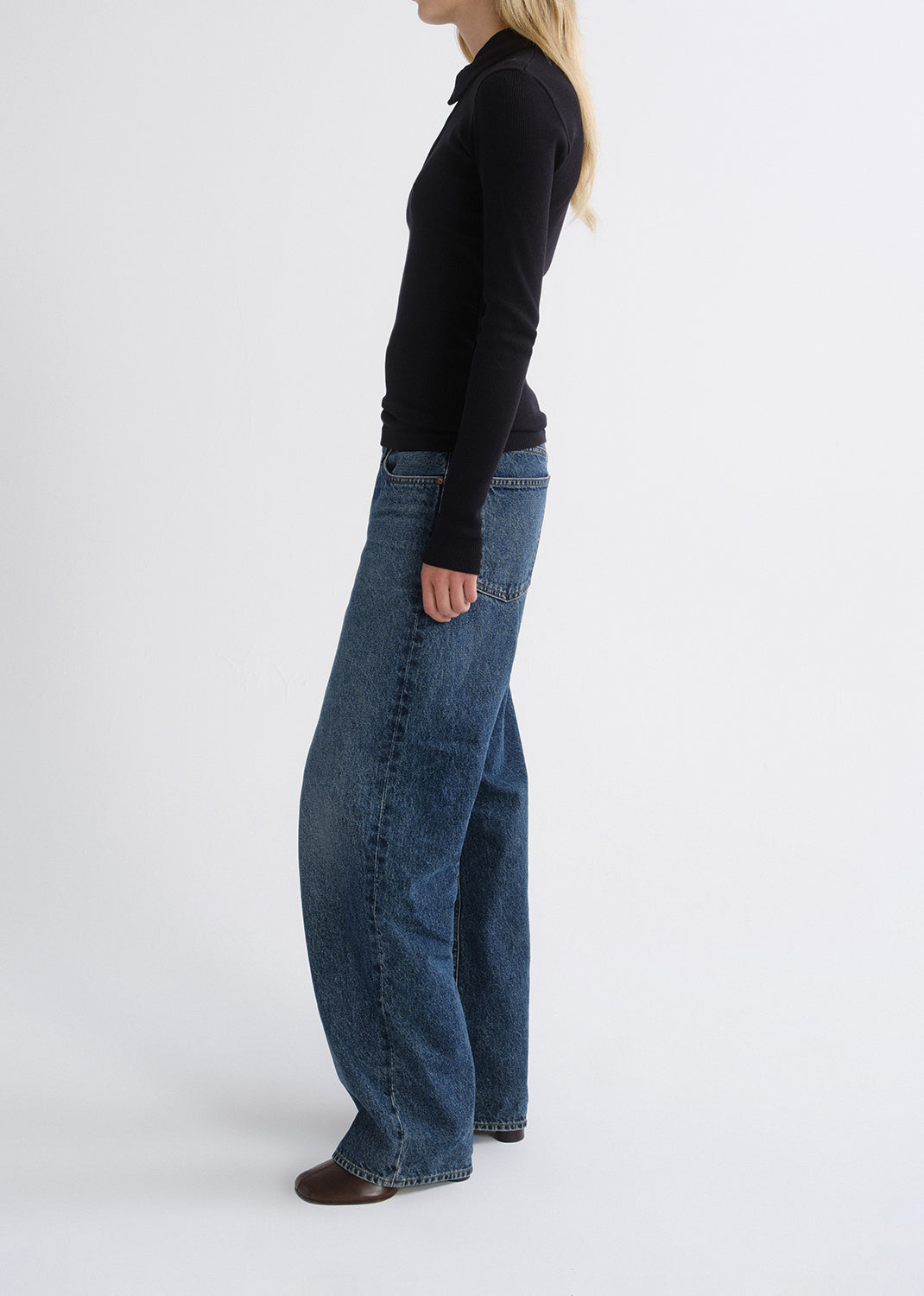 Person wearing a black fitted half-zip top with medium blue straight leg jeans viewed from the side on a grey background