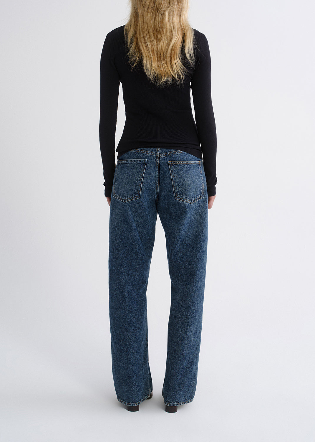 Person wearing a black fitted half-zip top with medium blue straight leg jeans viewed from the back on a grey background