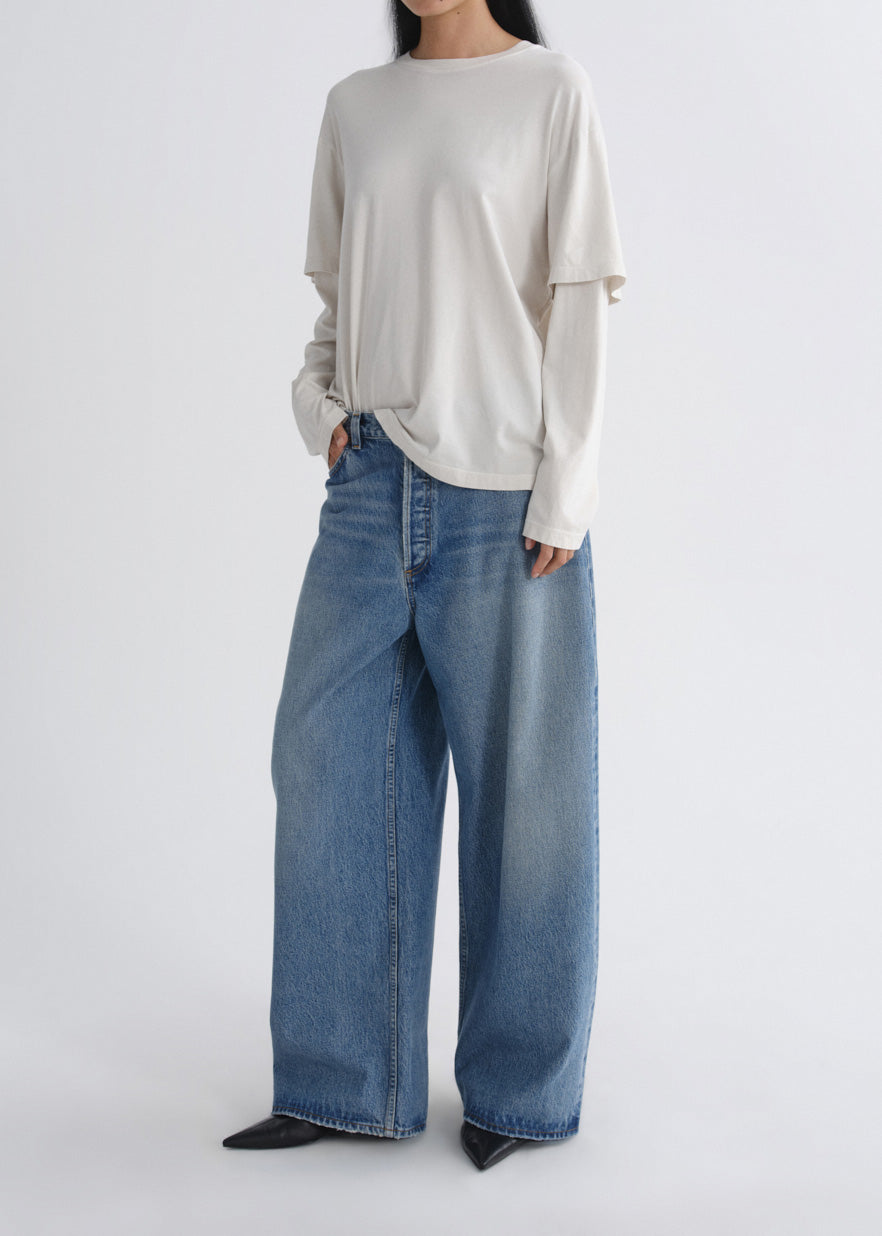 A model wearing a cream-colored long-sleeve top and wide-leg blue denim jeans, standing against a plain light gray studio background.
