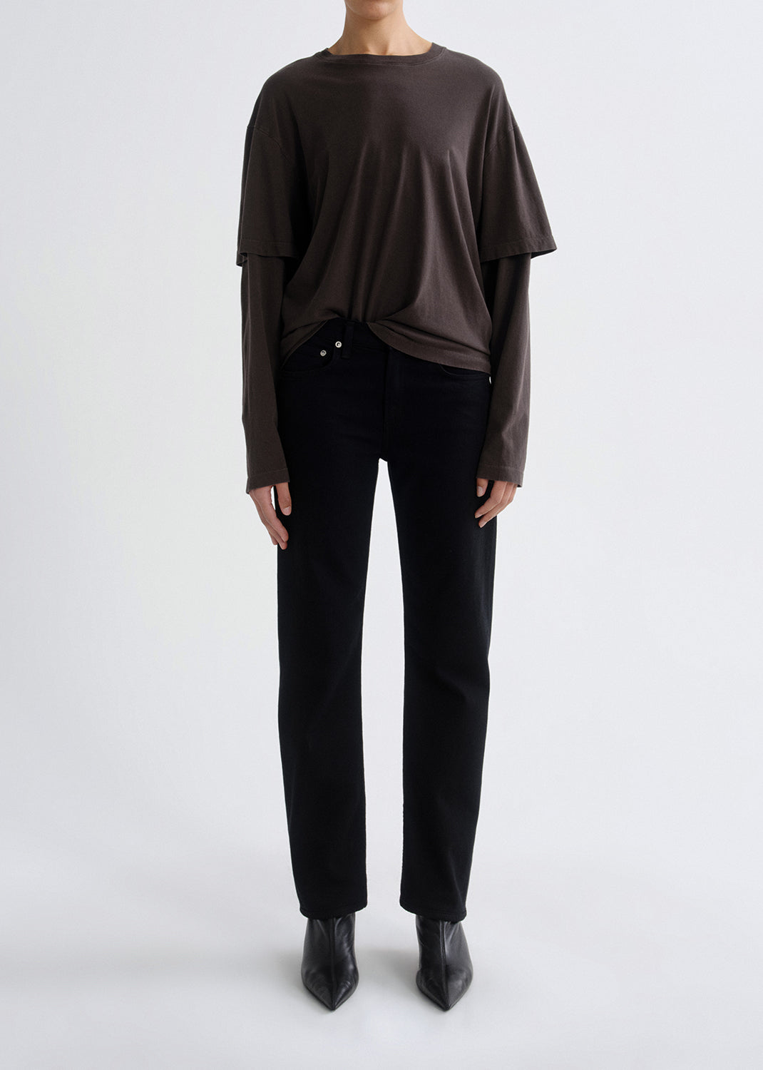 Person wearing a dark brown layered long sleeve top with black skinny jeans viewed from the back on a grey background.
