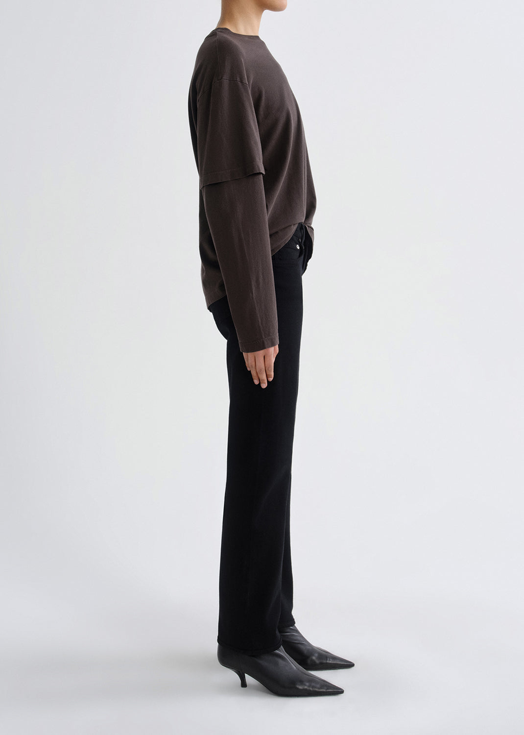 Person wearing a dark brown layered long sleeve top with black skinny jeans viewed from the side on a grey background.