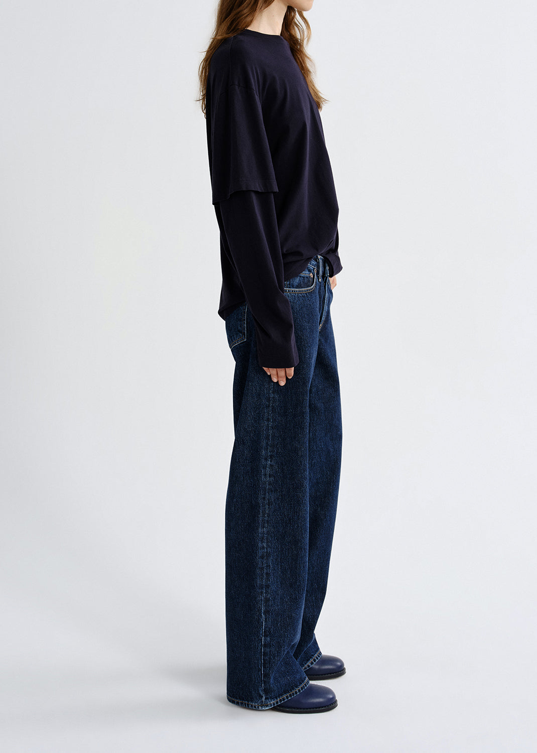 Person wearing a navy layered long sleeve top with dark wash wide leg jeans viewed from the side on a grey background.