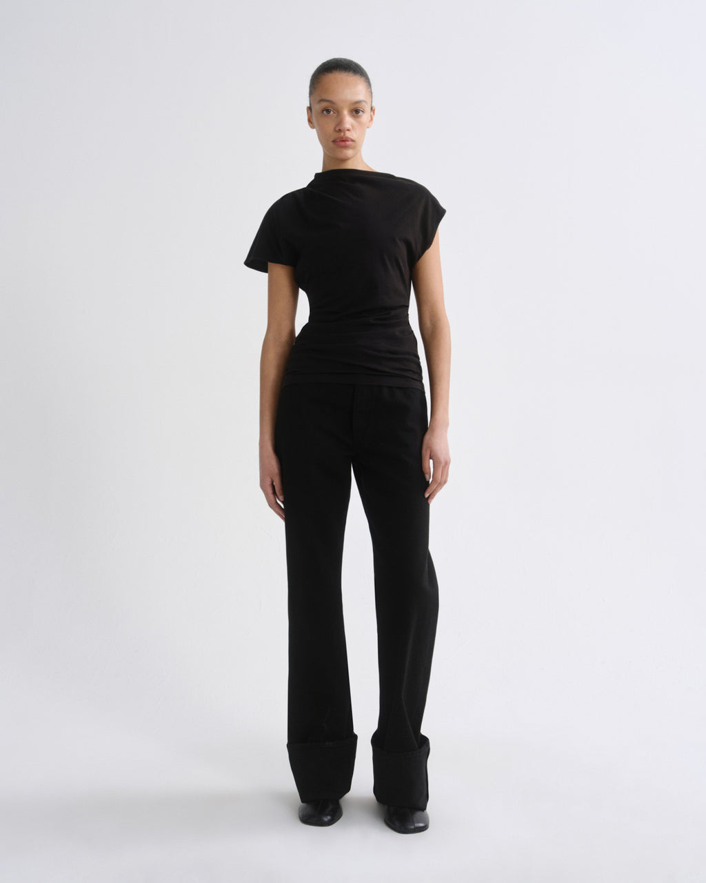 A model stands facing forward in a studio setting, wearing a black asymmetrical short-sleeve top and black wide-leg trousers against a light gray background.