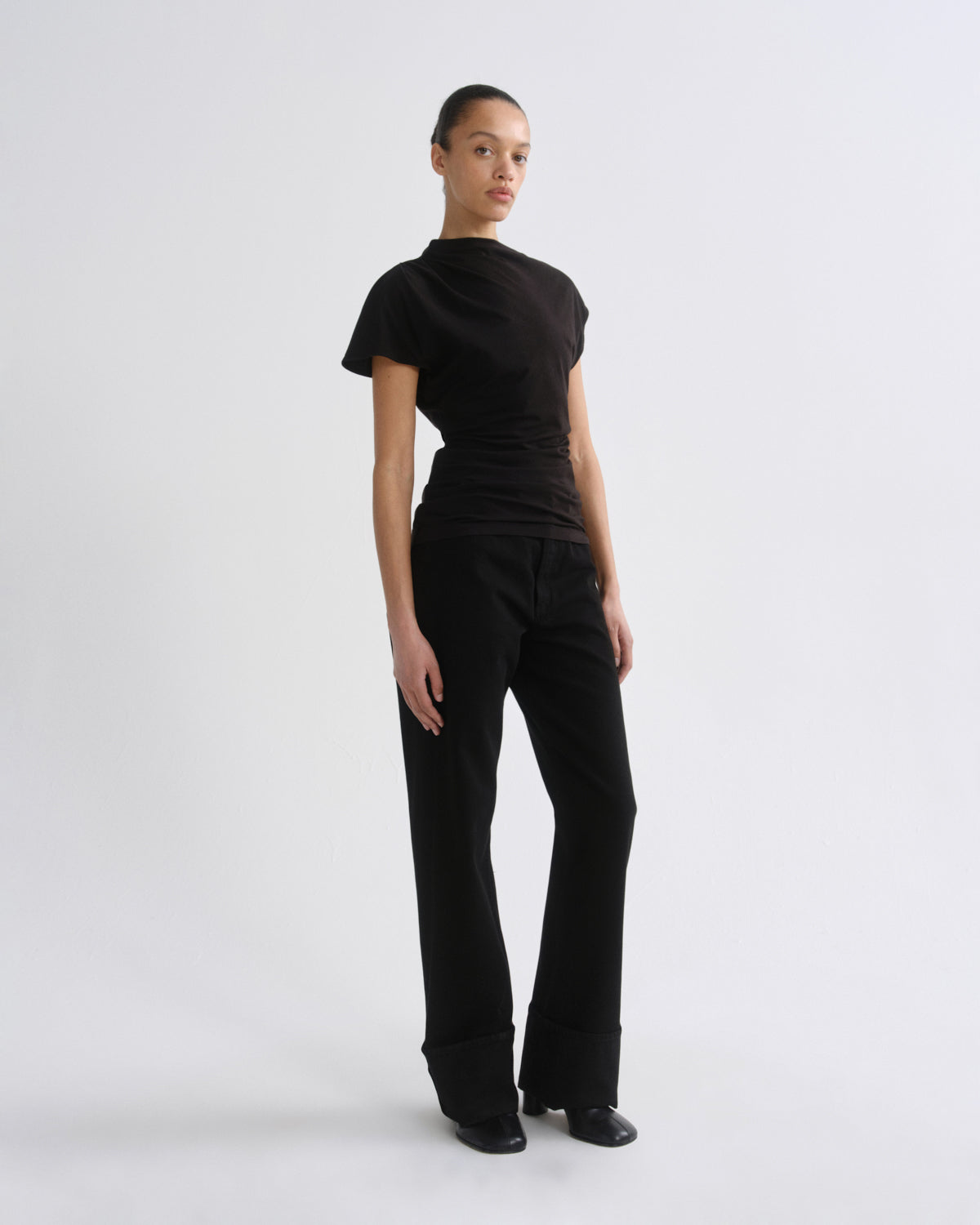 A model wearing a black short-sleeve mock neck top and matching black wide-leg trousers, standing against a light gray studio background.