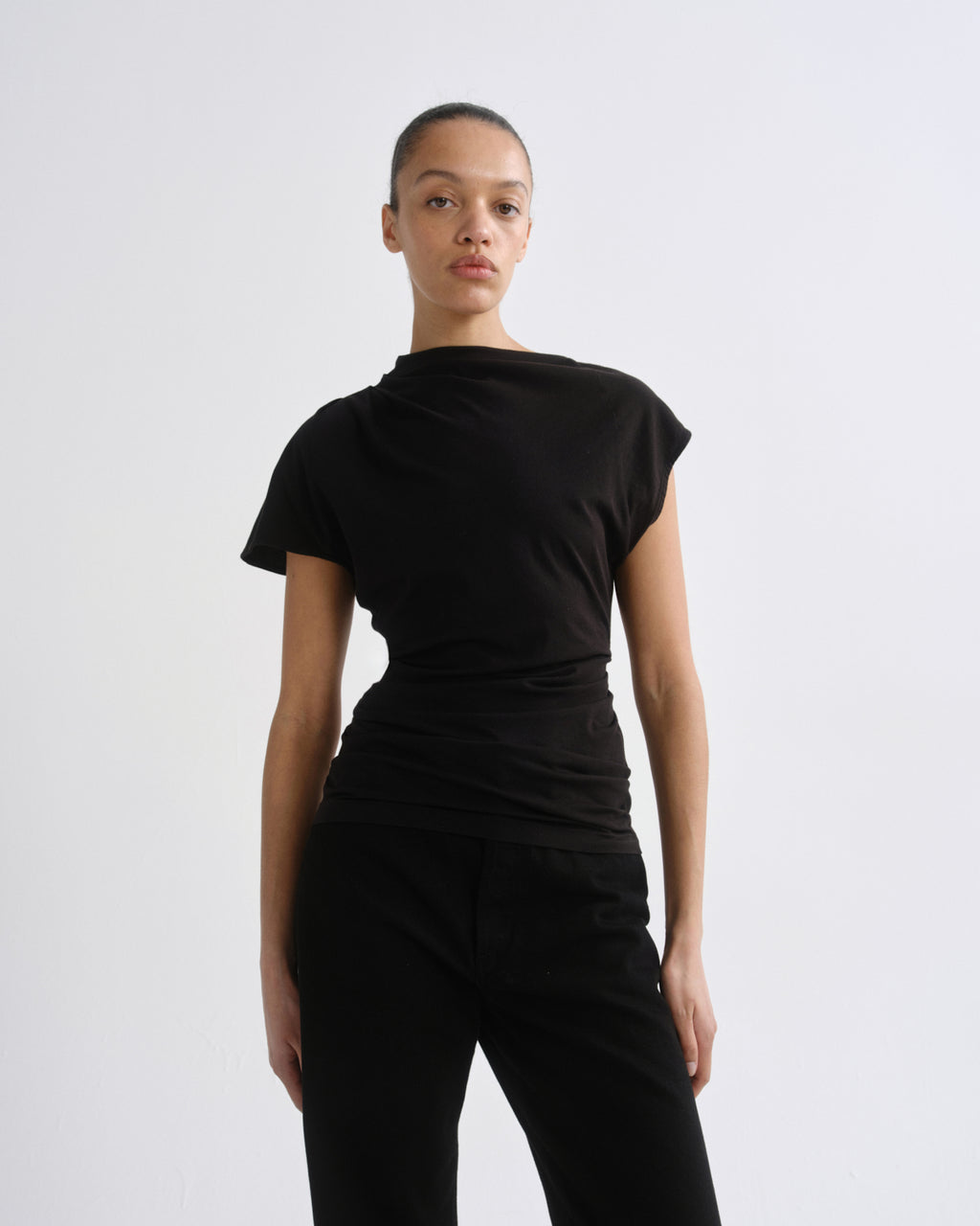 A model wearing a black short-sleeve top with a draped, asymmetrical neckline and black pants, photographed against a plain light gray studio background.